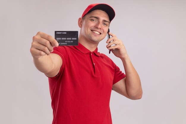 Smiling young delivery man wearing uniform with cap holding out credit card  speaks on phone isolated on white wall