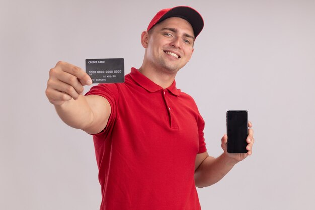 Smiling young delivery man wearing uniform with cap holding phone and holding out credit card at front isolated on white wall