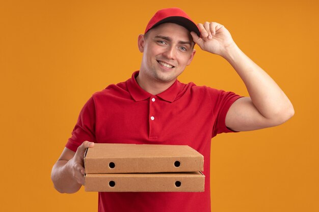 Smiling young delivery man wearing uniform with cap holding pizza boxes isolated on orange wall