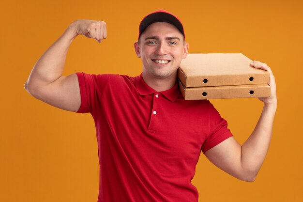 Smiling young delivery man wearing uniform with cap holding pizza boxes on shoulder and showing strong gesture isolated on orange wall