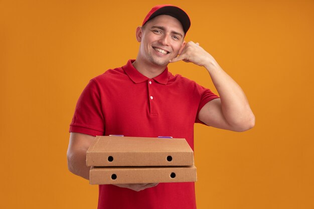 Smiling young delivery man wearing uniform with cap holding pizza boxes showing phone call gesture isolated on orange wall