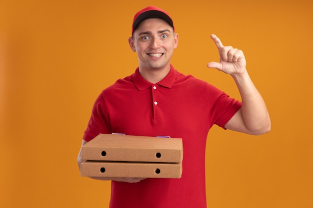 오렌지 벽에 고립 된 크기를 보여주는 피자 상자를 들고 모자와 유니폼을 입고 웃는 젊은 배달 남자