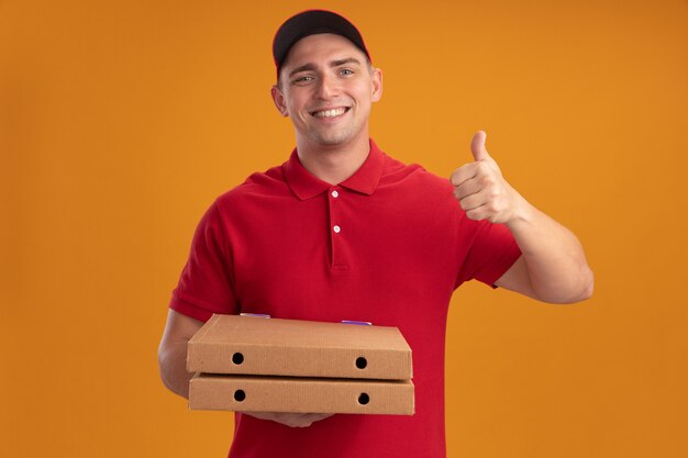 Smiling young delivery man wearing uniform with cap holding pizza boxes showing thumb up isolated on orange wall