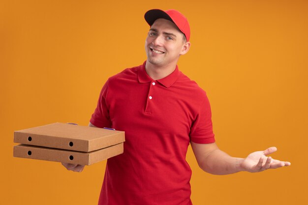 Улыбающийся молодой доставщик в униформе с кепкой держит коробки для пиццы, протягивая руку, изолированную на оранжевой стене