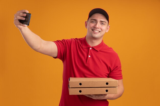 ピザの箱を保持している帽子と制服を着て笑顔の若い配達人はオレンジ色の壁に隔離されたselfieを取る