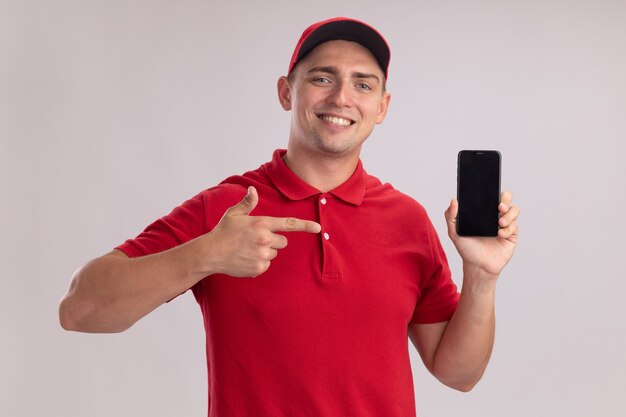 Улыбающийся молодой курьер в униформе с кепкой держит и указывает на телефон, изолированный на белой стене