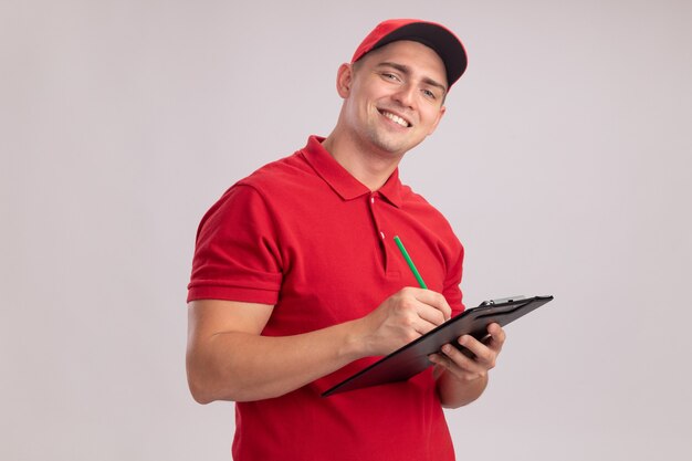 Smiling young delivery man wearing uniform with cap writing something in clipboard isolated on white wall
