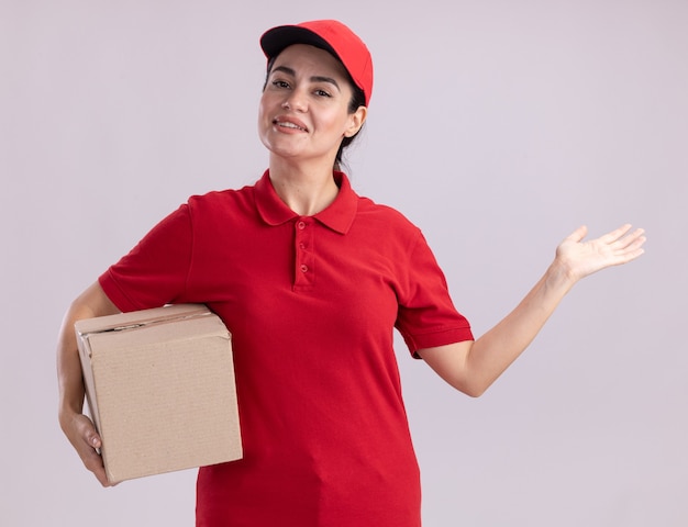 Улыбающаяся молодая женщина-доставщик в униформе и кепке держит картонную коробку с пустой рукой