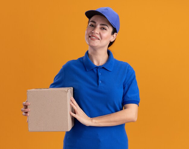 Улыбающаяся молодая женщина-доставщик в униформе и кепке держит картонную коробку, изолированную на оранжевой стене с копией пространства