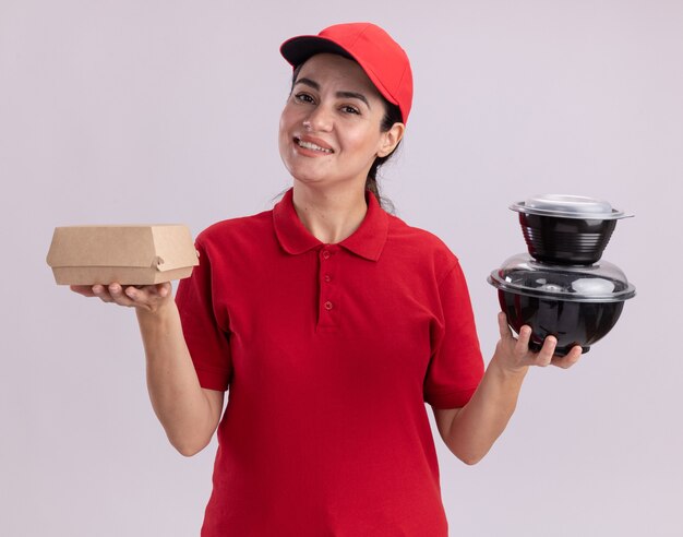 Smiling young delivery woman in uniform and cap holding paper food package and food containers  isolated on white wall