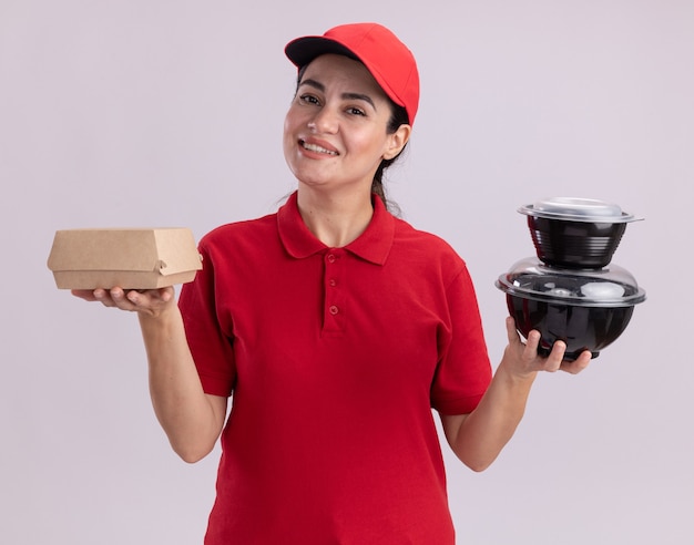 Улыбающаяся молодая женщина-доставщик в униформе и кепке, держащая бумажный пищевой пакет и пищевые контейнеры, изолированные на белой стене