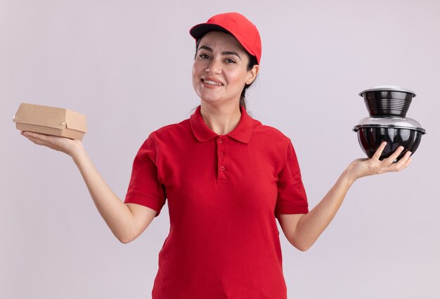 Smiling young delivery woman in uniform and cap holding paper food package and food containers looking at front isolated on white wall