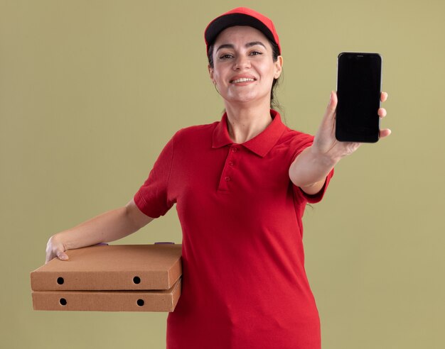 Улыбающаяся молодая женщина-доставщик в униформе и кепке держит упаковки с пиццей и протягивает мобильный телефон, изолированную на оливково-зеленой стене