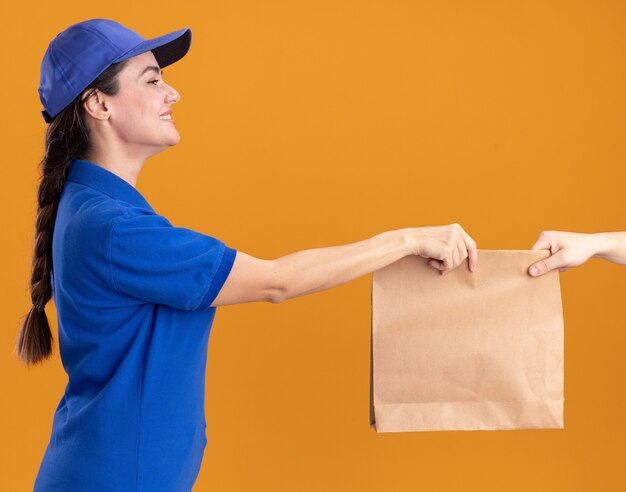 Smiling young delivery woman in uniform and cap standing in profile view giving paper package to client looking at client