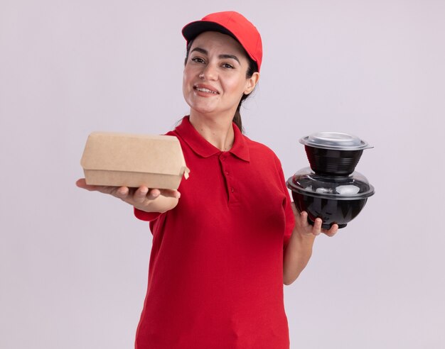 Улыбающаяся молодая женщина-доставщик в униформе и кепке, протягивающая бумажный пакет с едой и держащая пищевые контейнеры, глядя на перед, изолированную на белой стене