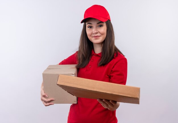 상자를 들고 빨간 모자에 빨간 티셔츠를 입고 웃는 젊은 배달 여자와 격리 된 흰 벽에 피자 상자를 보유