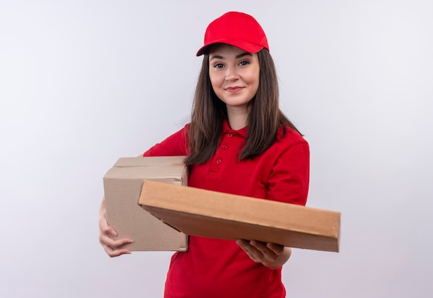 Smiling young delivery woman wearing red t-shirt in red cap holding a box and holds out the pizza box on isolated white wall