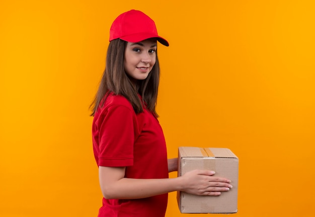 Sorridente giovane donna di consegna che indossa la maglietta rossa in berretto rosso che tiene una scatola sulla parete gialla isolata