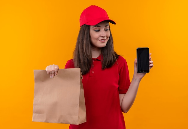 Улыбающаяся молодая женщина-доставщик в красной футболке в красной кепке держит пакет и телефон на изолированной желтой стене