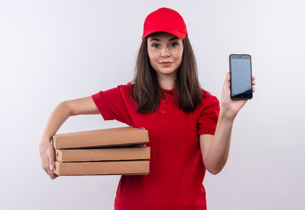 Улыбающаяся молодая женщина-доставщик в красной футболке в красной кепке держит коробку для пиццы и телефон на изолированной белой стене