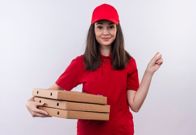 Улыбающаяся молодая женщина-доставщик в красной футболке в красной кепке держит коробку для пиццы и указывает на изолированную белую стену