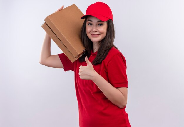 Smiling young delivery woman wearing red t-shirt in red cap holding pizza box and showing thumbs up on isolated white wall