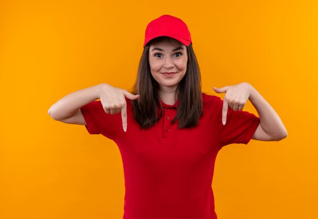 赤い帽子に赤いtシャツを着ている若い配達の女性の笑顔と孤立したオレンジ色の壁に指を下向き