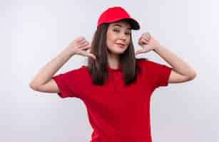 Free photo smiling young delivery woman wearing red t-shirt in red cap points to himself on isolated white wall