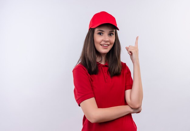 Улыбающаяся молодая женщина-доставщик в красной футболке в красной кепке указывает вверх на изолированной белой стене