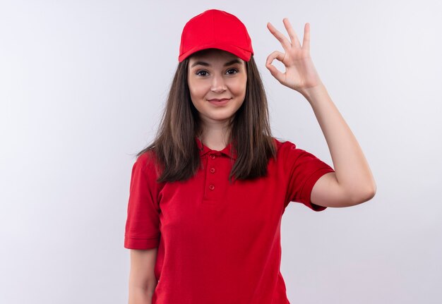 Smiling young delivery woman wearing red t-shirt in red cap shows okay gesture on isolated white wall