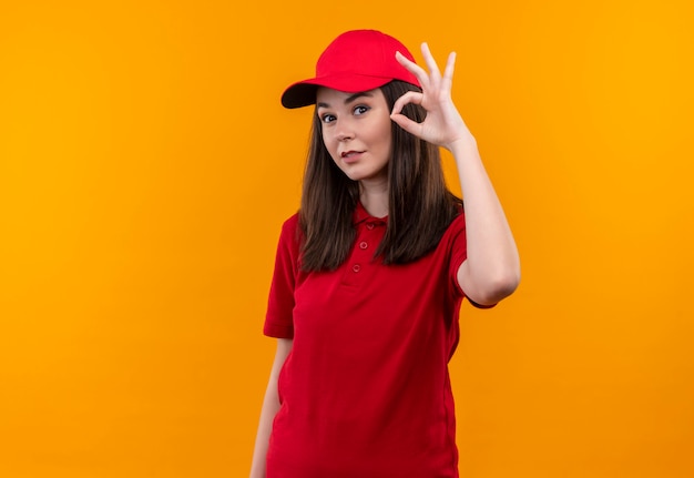 빨간 모자에 빨간 티셔츠를 입고 웃는 젊은 배달 여자와 격리 된 노란색 벽에 좋아요 gasture를 보여줍니다
