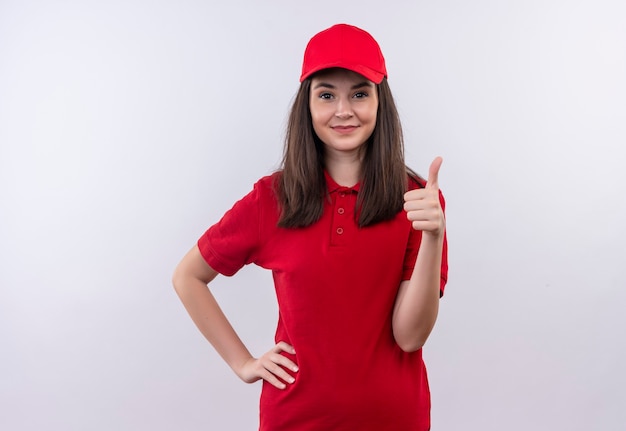 Улыбающаяся молодая женщина-доставщик в красной футболке в красной кепке показывает палец вверх на изолированной белой стене