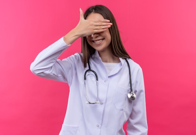 Smiling young doctor girl wearing stethoscope medical gown covered eyes with hand on isolated pink background
