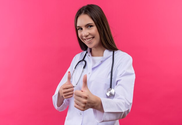 Улыбающаяся молодая девушка-врач в медицинском халате со стетоскопом показывает палец вверх на изолированном розовом фоне