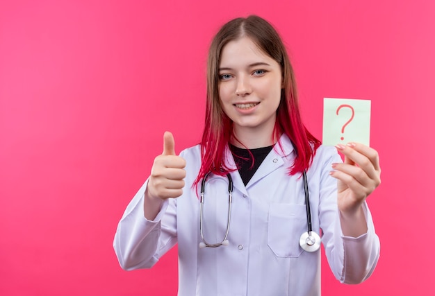 Smiling young doctor girl wearing stethoscope medical gown holding paper question mark her thumb up on pink isolated background