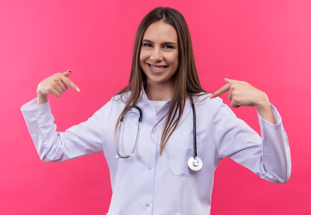Smiling young doctor girl wearing stethoscope medical gown points to herself on isolated pink background