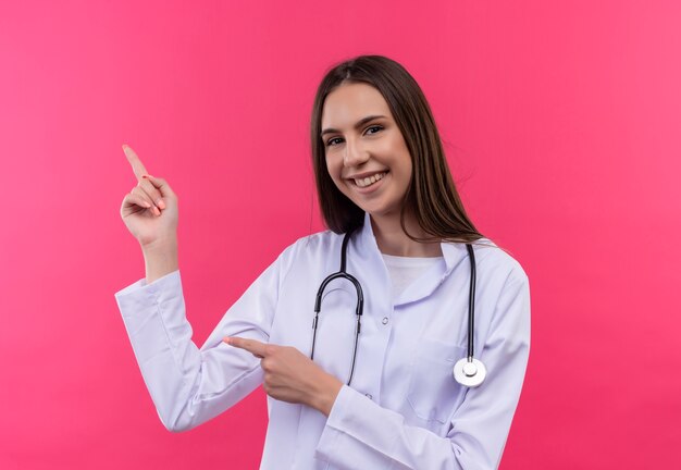 Smiling young doctor girl wearing stethoscope medical gown points to side on isolated pink background