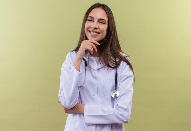 緑の背景のあごに手を置いて聴診器の医療用ガウンを着て笑顔の若い医者の女の子