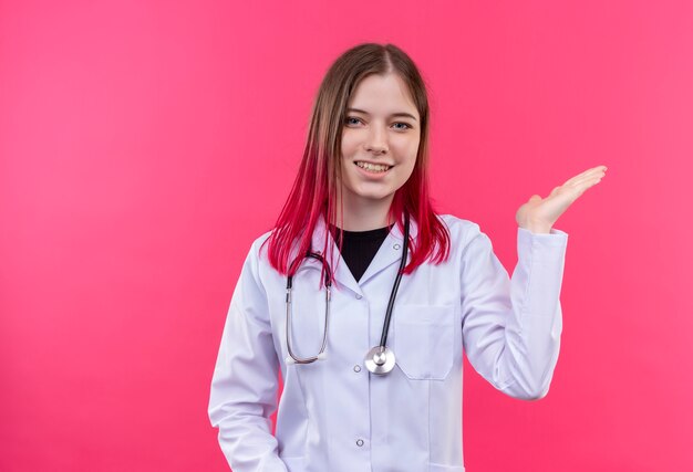 ピンクの孤立した背景に手を上げる聴診器医療用ガウンを着て笑顔の若い医者の女の子