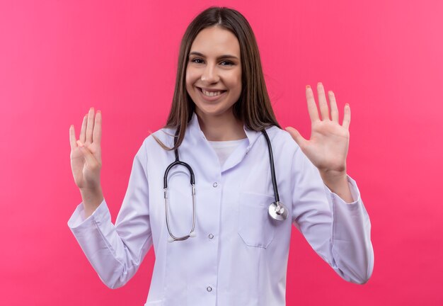 Smiling young doctor girl wearing stethoscope medical gown showing different numbers on isolated pink background