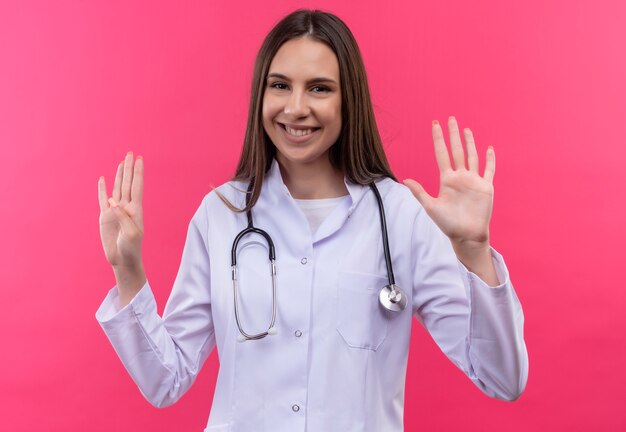 孤立したピンクの背景に異なる数を示す聴診器の医療用ガウンを着て笑顔の若い医者の女の子