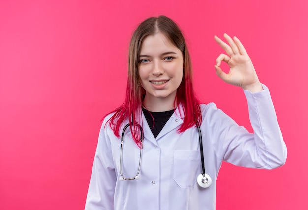 분홍색 격리 된 배경에 좋아요 제스처를 보여주는 청진 기 의료 가운을 입고 웃는 젊은 의사 소녀