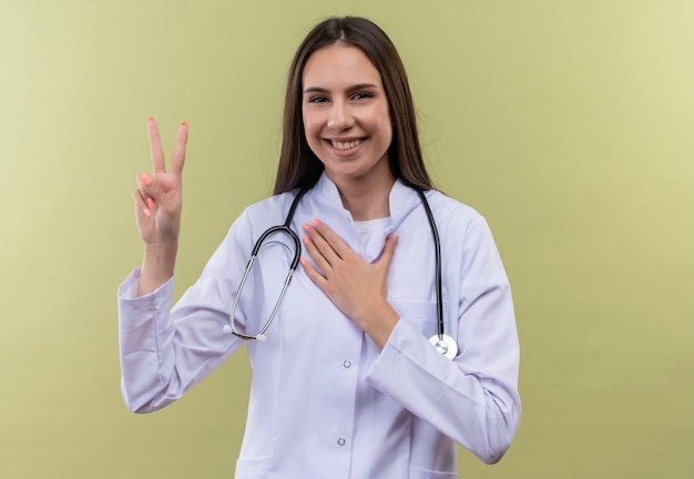 平和のジェスチャーを示す聴診器の医療用ガウンを着て笑顔の若い医者の女の子は、緑の背景に彼女の手を心に置きます