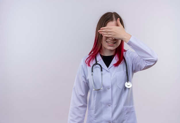 Улыбающаяся молодая девушка-врач в медицинском халате со стетоскопом закрыла глаза рукой на изолированном белом фоне с копией пространства
