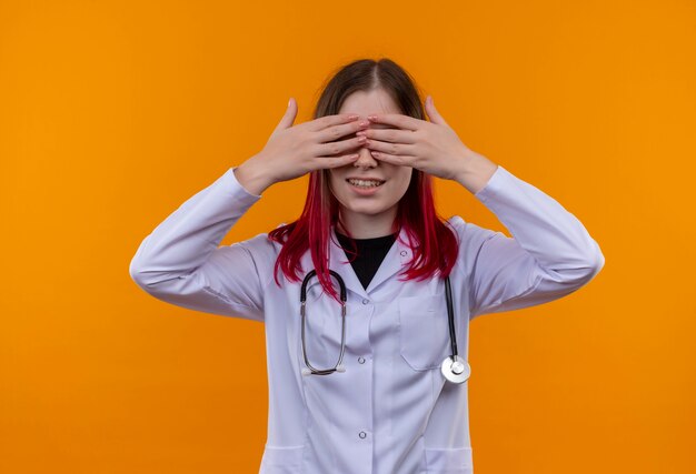 Smiling young doctor girl wearing stethoscope medical robe covered eyes with hands on isolated orange background