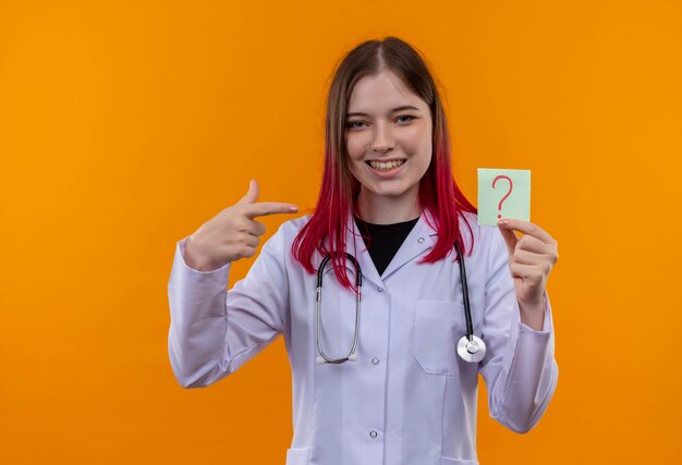 Улыбающаяся молодая девушка-врач в медицинском халате со стетоскопом указывает пальцем на бумажный вопросительный знак на руке на изолированном оранжевом фоне