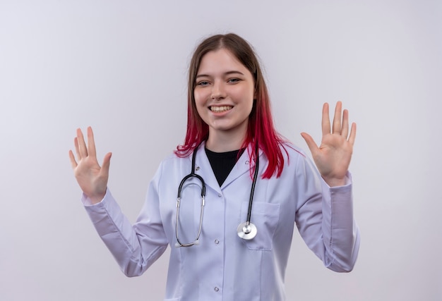 격리 된 흰색 배경에 손을 올리는 청진 기 의료 가운을 입고 웃는 젊은 의사 소녀