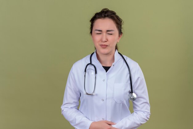 Smiling young doctor wearing medical gown wearing stethoscope closed eyes on green wall