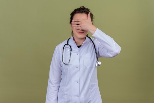 Smiling young doctor wearing medical gown wearing stethoscope covered eyes on green wall