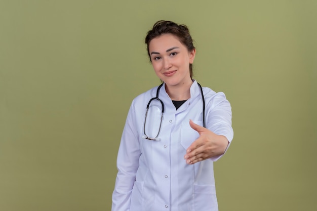 Smiling young doctor wearing medical gown wearing stethoscope held out her hand on green wall